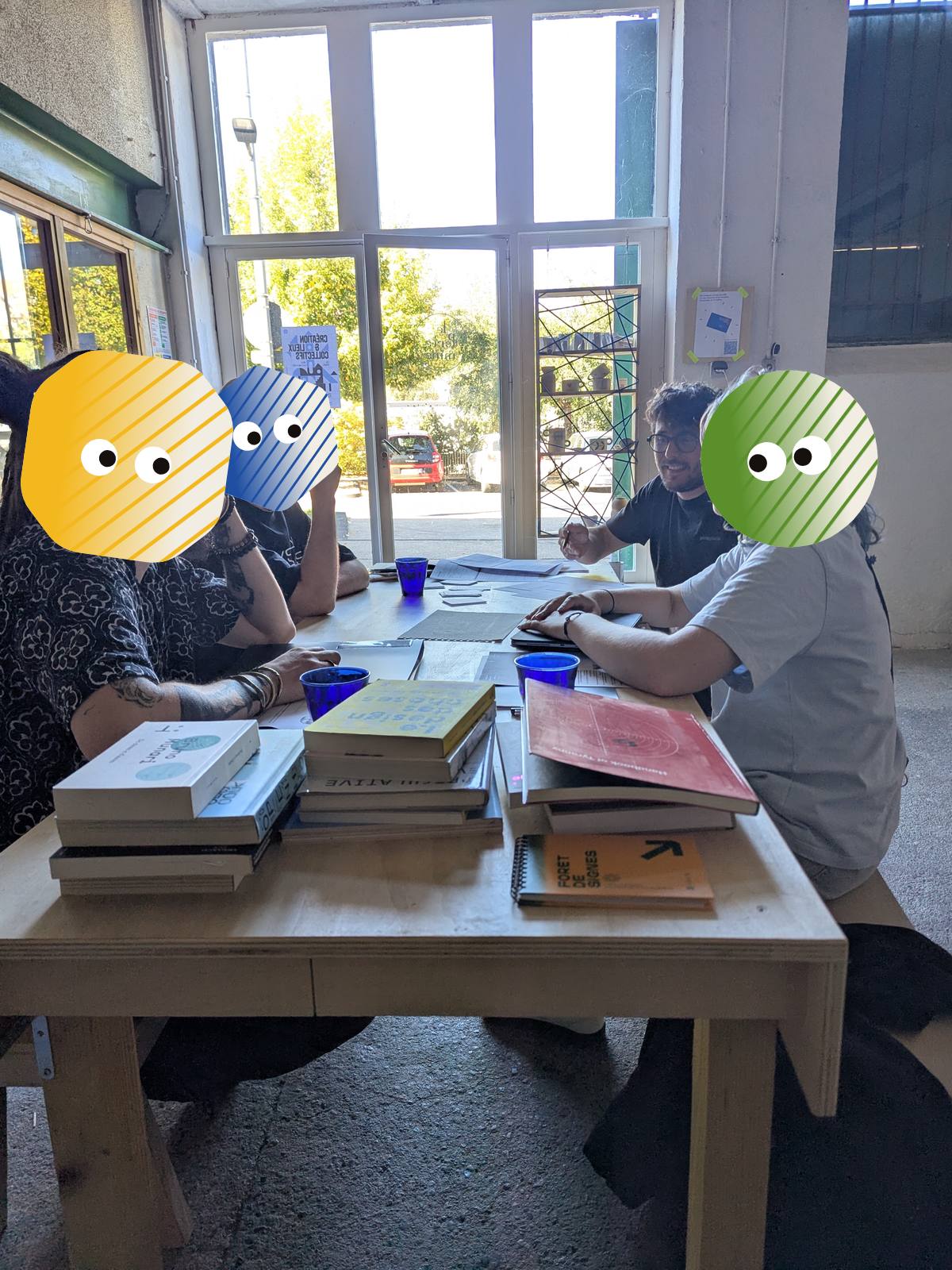 Jonathan Mazuel en discussion avec trois participants autour de la table couverte de livres. Les visages des participants sont masqués par des personnages colorés.